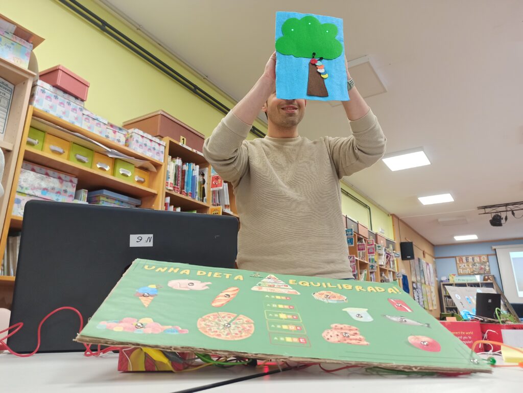 Imagen de un docente aguantando un proyecto de e-textil mientras en la mesa hay otro realizado con Makey Makey. Todos ellos sobre alimentación saludable